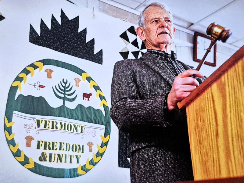 An elderly man stands at a podium holding a gavel, with the Vermont state seal and the words "Vermont Freedom & Unity" displayed on a banner behind him.
