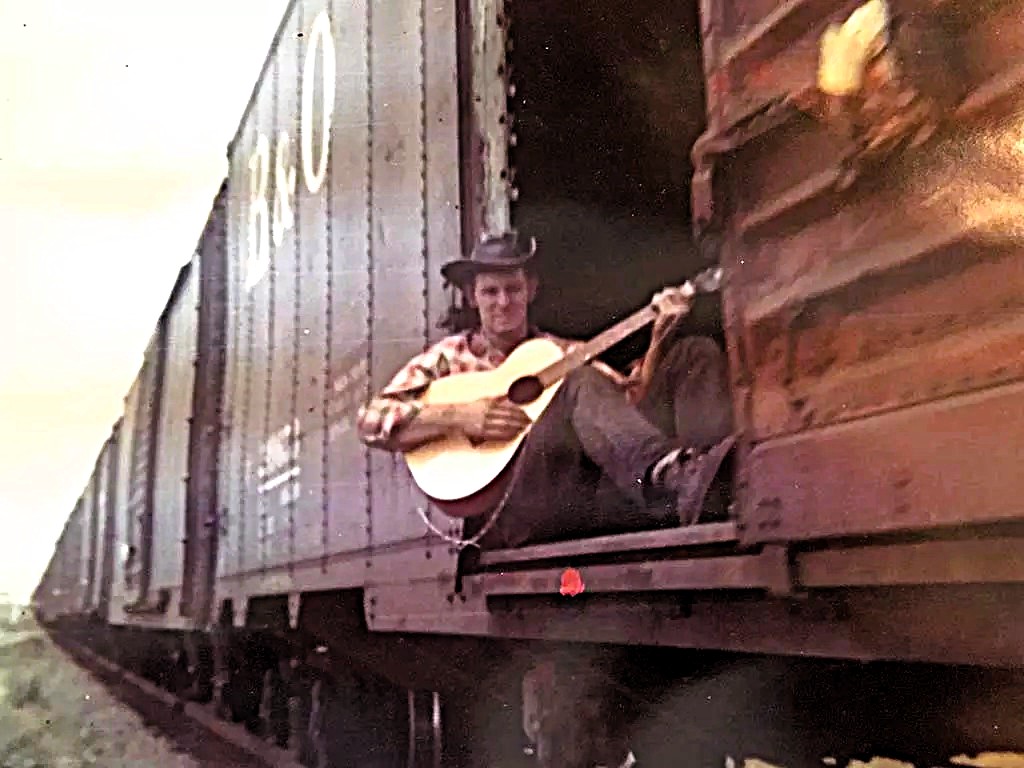 A person sits on the open doorway of a moving boxcar, playing an acoustic guitar, with several train cars visible in the background.
