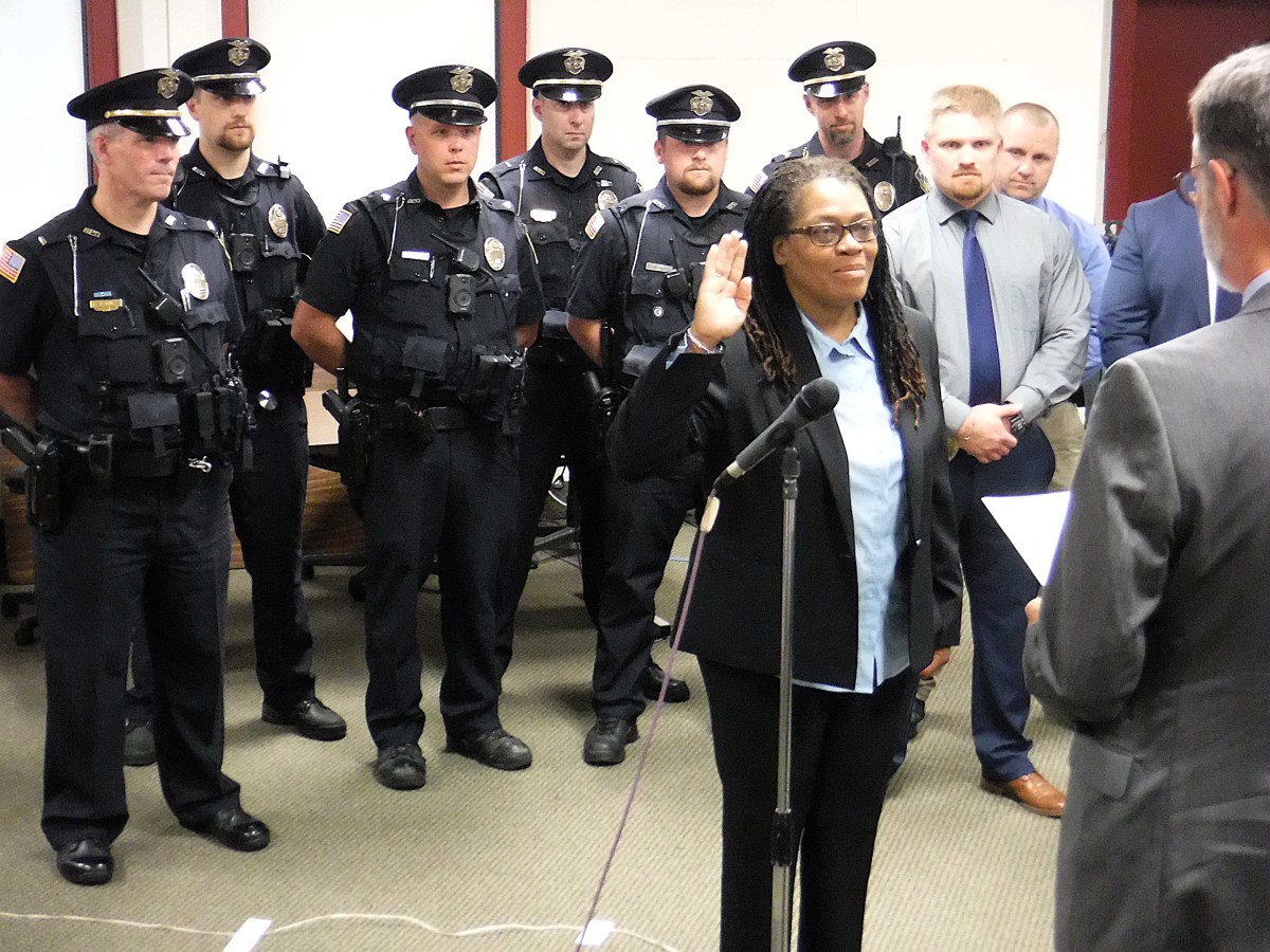 A woman raises her right hand to take an oath in front of a man holding a paper, while several uniformed police officers and others stand in the background.