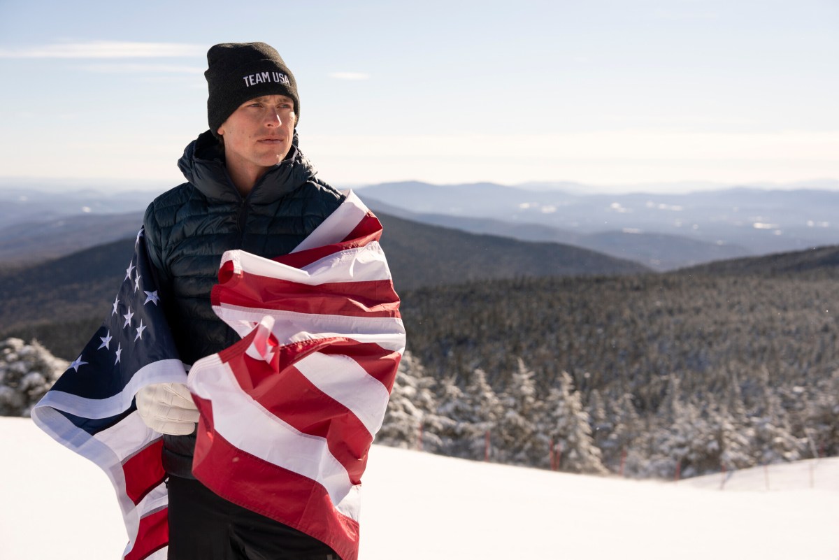 Person wearing a "Team USA" hat stands on a snowy mountain slope, wrapped in an American flag, with forested hills and a clear sky in the background.