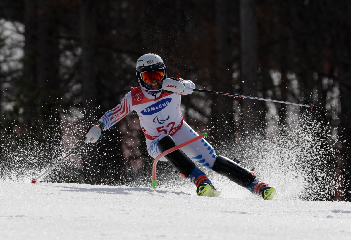 A para-alpine skier wearing a helmet and bib number 52 navigates a turn on a snowy slope, with snow spraying from their skis.