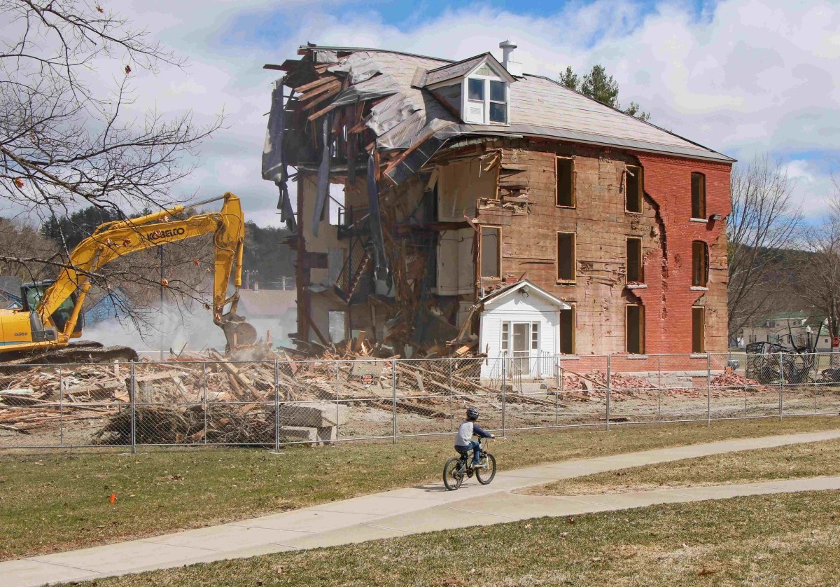 A partially demolished brick building stands behind a fence as an excavator works nearby; a child rides a bicycle on a path in the foreground.