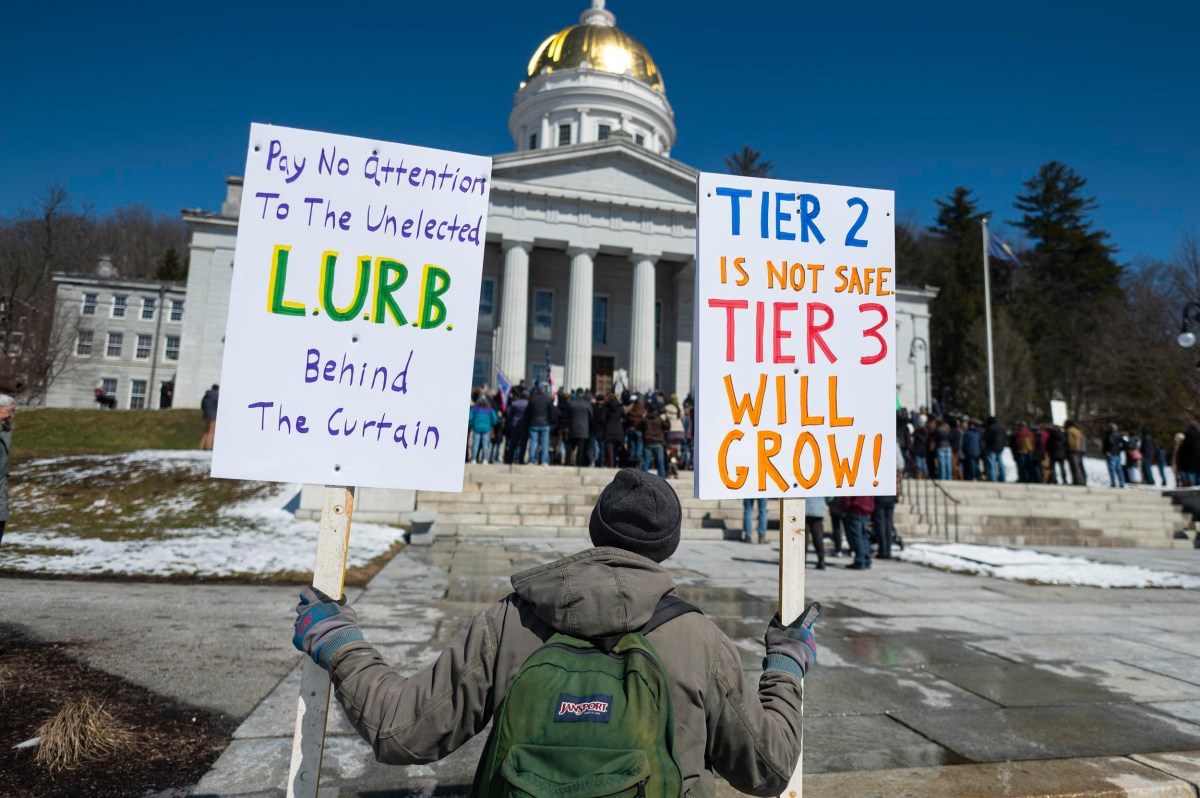 A person stands in front of a government building holding two protest signs about L.U.R.B. and concerns with Tier 2 and Tier 3 policies, as a crowd gathers outside.