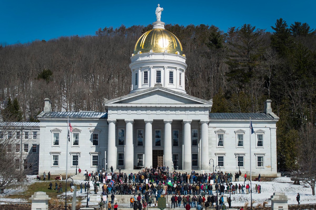 People gather outside a large white building with a gold-domed roof, columns, and statues, surrounded by trees and patches of snow.
