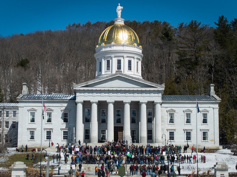 People gather outside a large white building with a gold-domed roof, columns, and statues, surrounded by trees and patches of snow.