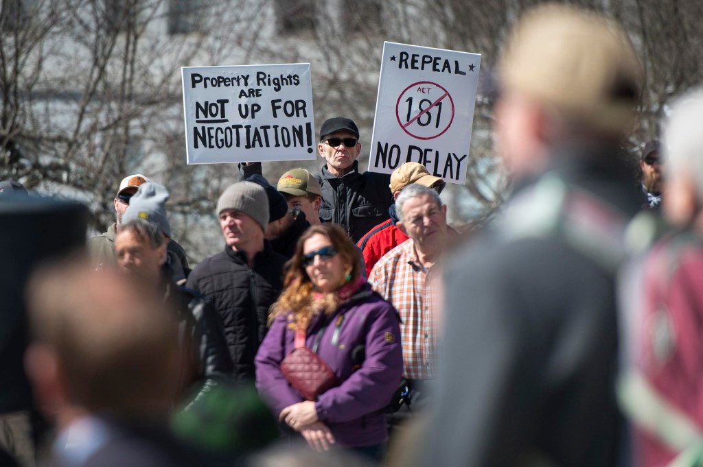 A group of people gathers outdoors; two prominent signs read "Property rights are not up for negotiation" and "Repeal Act 181 No delay.