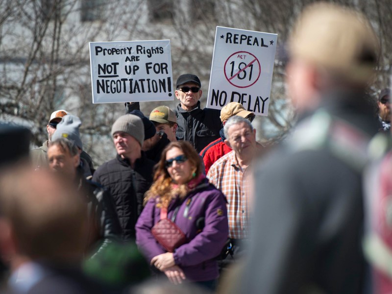 A group of people gathers outdoors; two prominent signs read "Property rights are not up for negotiation" and "Repeal Act 181 No delay.