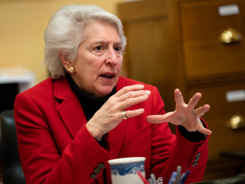 An older woman in a red blazer gestures with her hands while speaking, seated at a desk with a cup and office supplies in front of her.