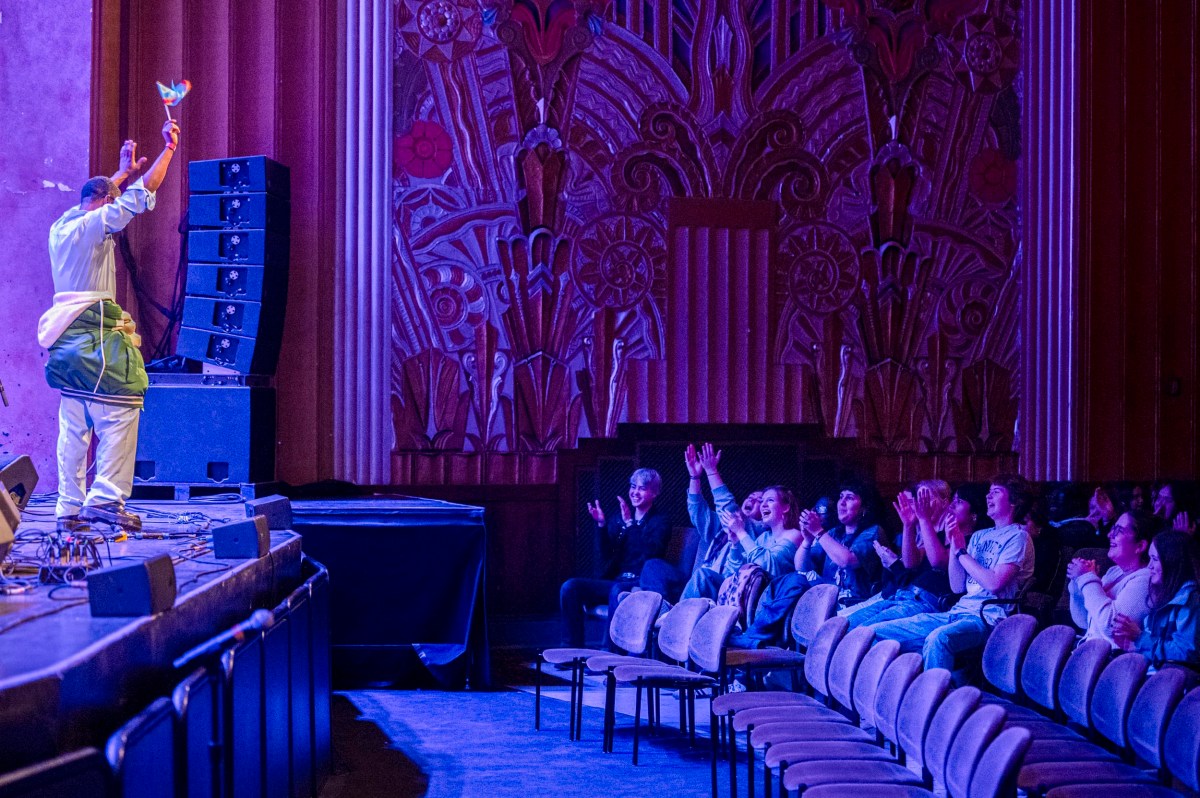 A performer on stage raises their hand to a small audience who are clapping in an ornate, mostly empty theater with dramatic lighting.