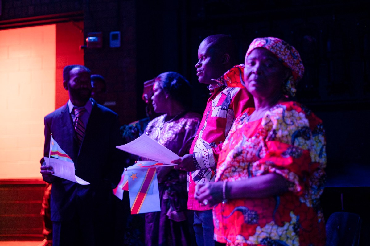 Four people stand indoors holding papers and Democratic Republic of the Congo flags, dressed in colorful attire, with purple and red lighting.