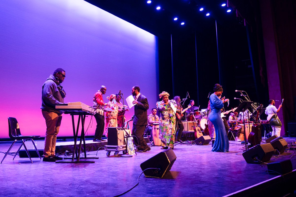 A band performs on stage under purple and blue lighting, with musicians playing instruments and singers in colorful outfits.