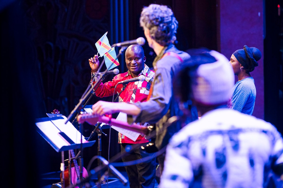 A musician on stage holds up a small Democratic Republic of the Congo flag, surrounded by other performers, with music stands and instruments visible.