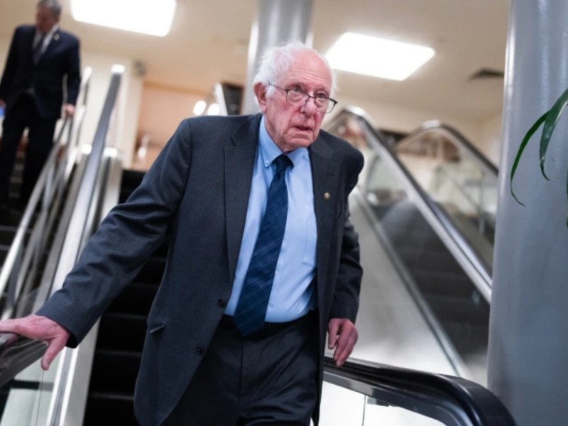 An older man in a suit and tie rides an escalator indoors, with another person in the background near a staircase.