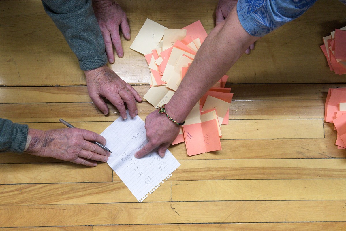 Two people work at a wooden table, one writing on a sheet of paper while the other points, with stacks of colored sticky notes beside them.