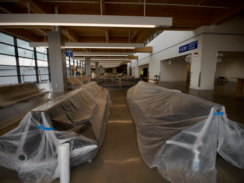 Rows of airport seating covered in plastic sheeting, situated in a modern, empty terminal area with large windows and overhead signs.