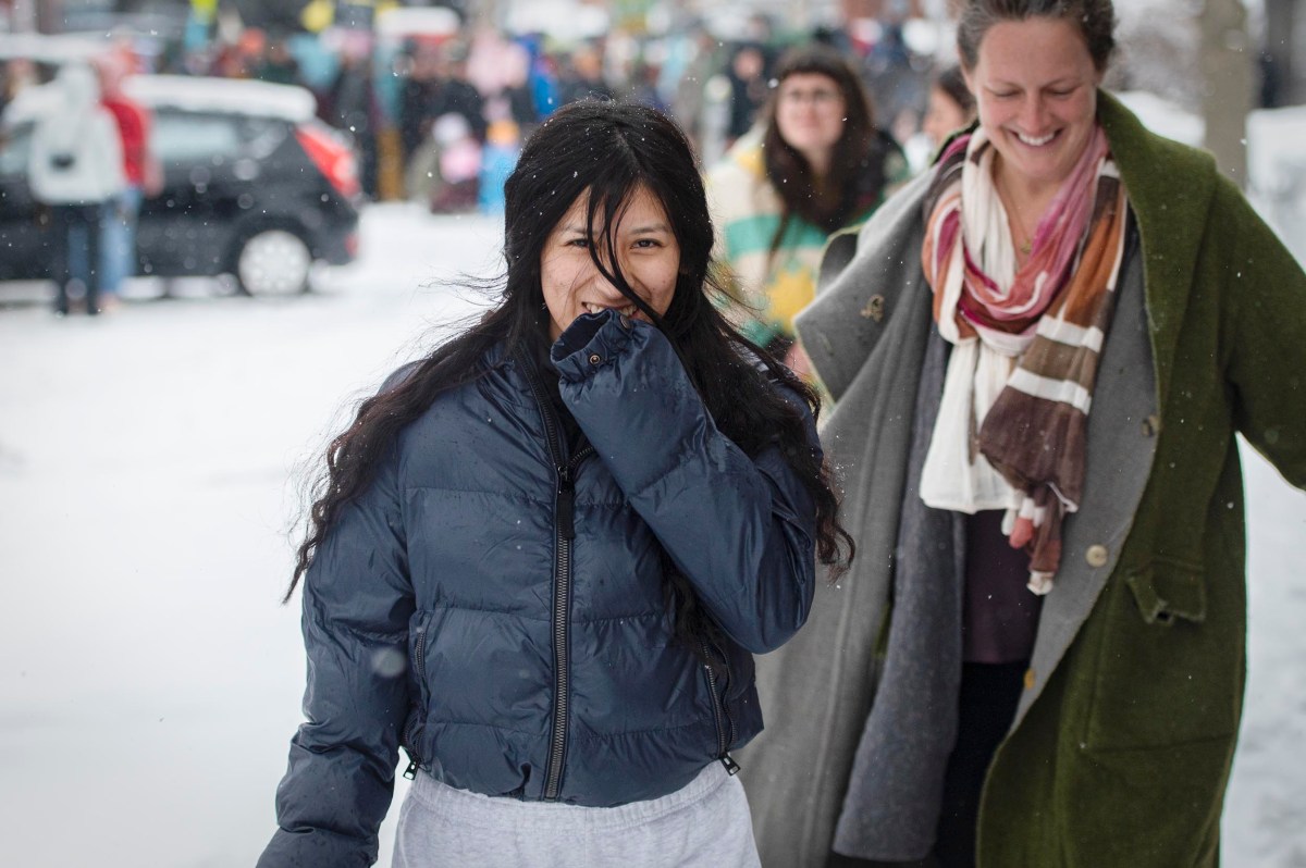 Two people walk outside on a snowy day. One person in front wears a dark jacket and covers their mouth; the other behind wears a scarf and smiles. Other people and cars are in the background.