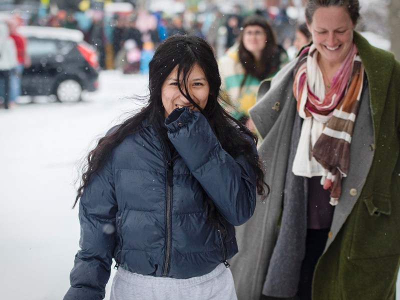 Two people walk outside on a snowy day. One person in front wears a dark jacket and covers their mouth; the other behind wears a scarf and smiles. Other people and cars are in the background.