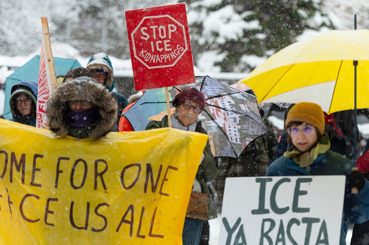 A group of people protest in snowy weather holding signs, including "STOP ICE KIDNAPPINGS," "ICE YA BASTA," and a large yellow banner. Some use umbrellas.