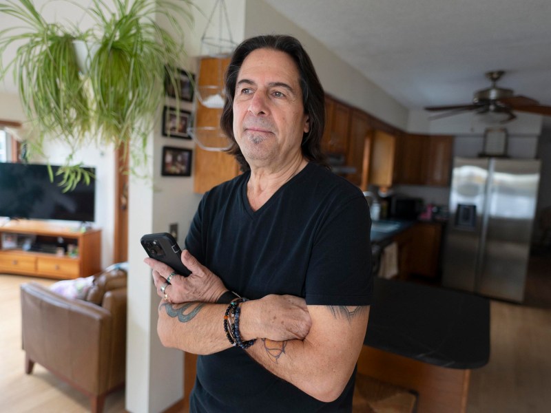 A man with long dark hair, wearing a black shirt, stands in a kitchen holding a smartphone, with his arms crossed. Houseplants and furniture are visible in the background.