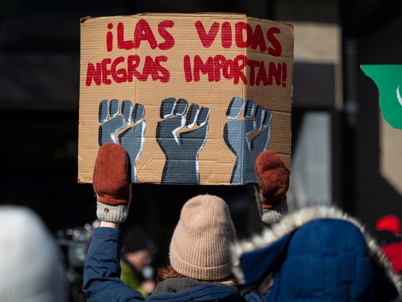 A person in winter clothing holds a cardboard sign reading "¡LAS VIDAS NEGRAS IMPORTAN!" with three raised fists painted below the text.
