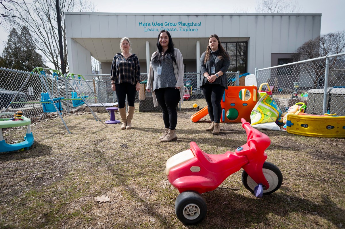 Three women stand in a fenced outdoor play area with various colorful toys in front of a building labeled "Here Wee Grow Playschool.