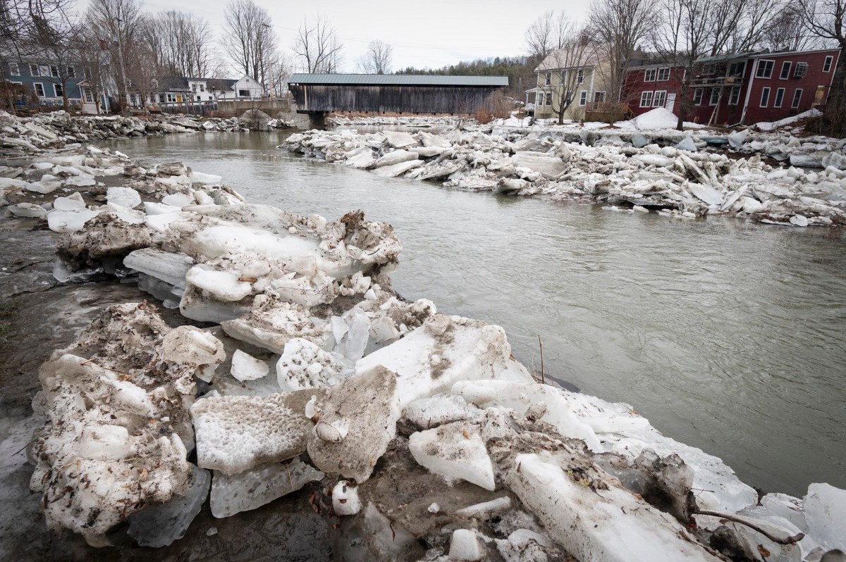 Large chunks of ice line the riverbank in front of a covered wooden bridge and houses on a cloudy winter day.