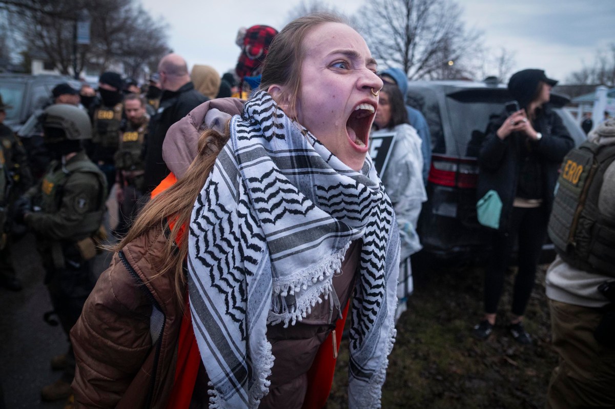 A person wearing a patterned scarf and orange vest shouts amid a crowd of people, some in tactical gear, during an outdoor protest or demonstration.