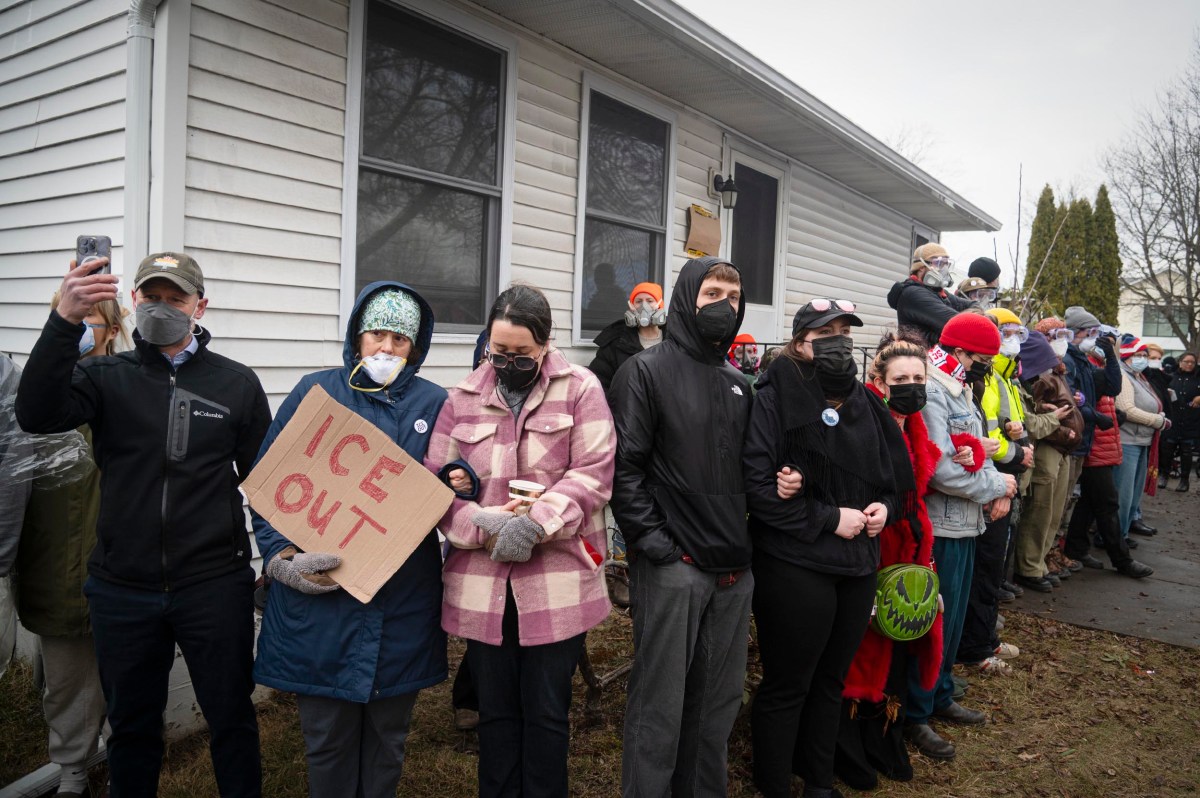 A group of people wearing winter clothes and face masks stand arm-in-arm along a building; one person holds a cardboard sign reading "ICE OUT.