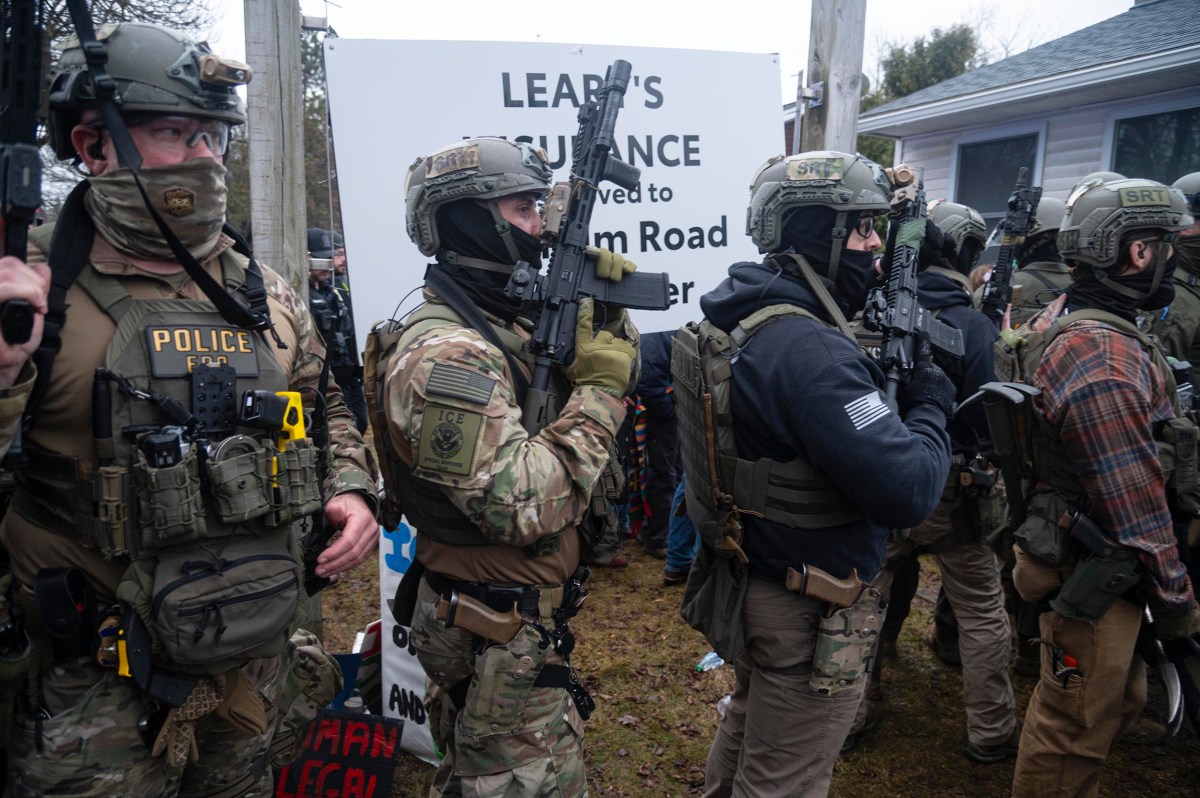 A group of heavily armed police officers in tactical gear stand in formation outdoors near a sign and a crowd.