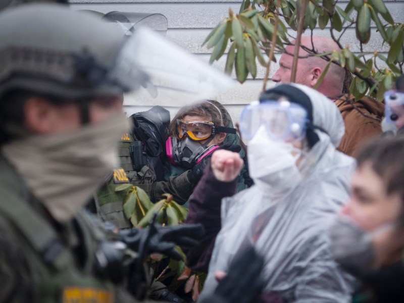 Law enforcement officers in riot gear face protesters wearing masks and goggles. Some protesters wear plastic coverings and appear to be shouting or gesturing.