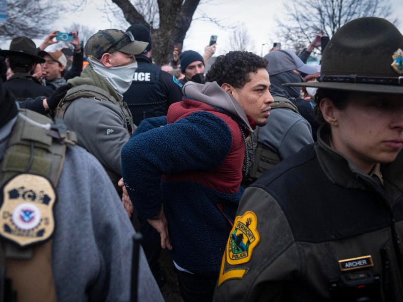 A man is handcuffed and escorted by police officers during a public event as bystanders take photos and videos.