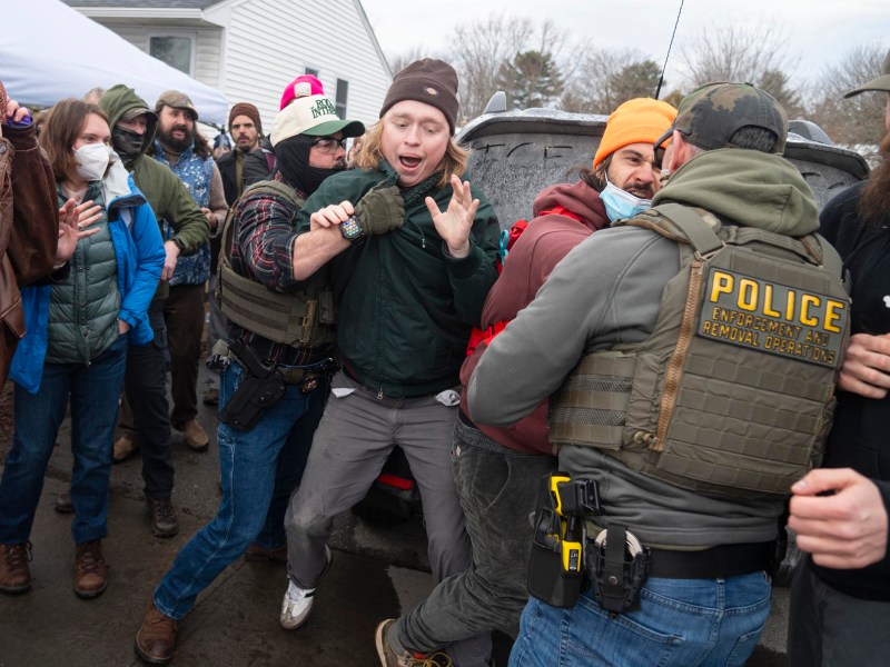 A group of law enforcement officers detain a man in a crowd outdoors; bystanders stand nearby, some wearing masks and winter clothing.