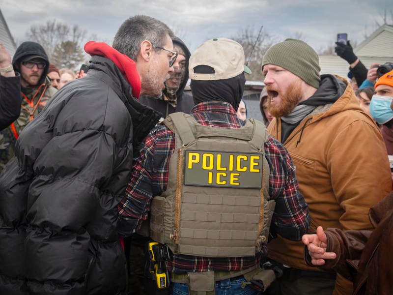 A person wearing a "Police ICE" vest stands between two men having a heated exchange in a crowd outdoors, with several onlookers in the background.
