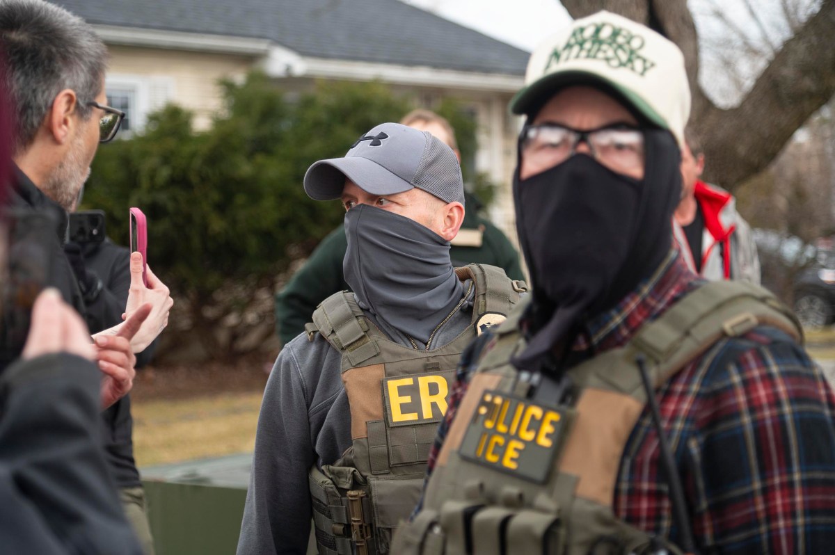 Two people wearing tactical vests labeled "POLICE ICE" and "ERS" stand outdoors, one wearing a mask and hat, as another person gestures with a smartphone nearby.