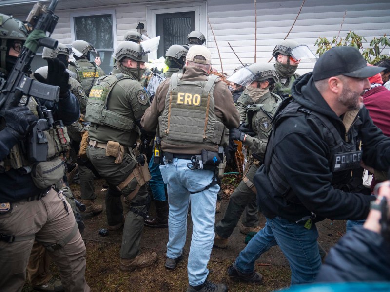 Law enforcement officers in tactical gear gather near the entrance of a house during a daytime operation. Some officers have "ERO" and "POLICE" vests visible.