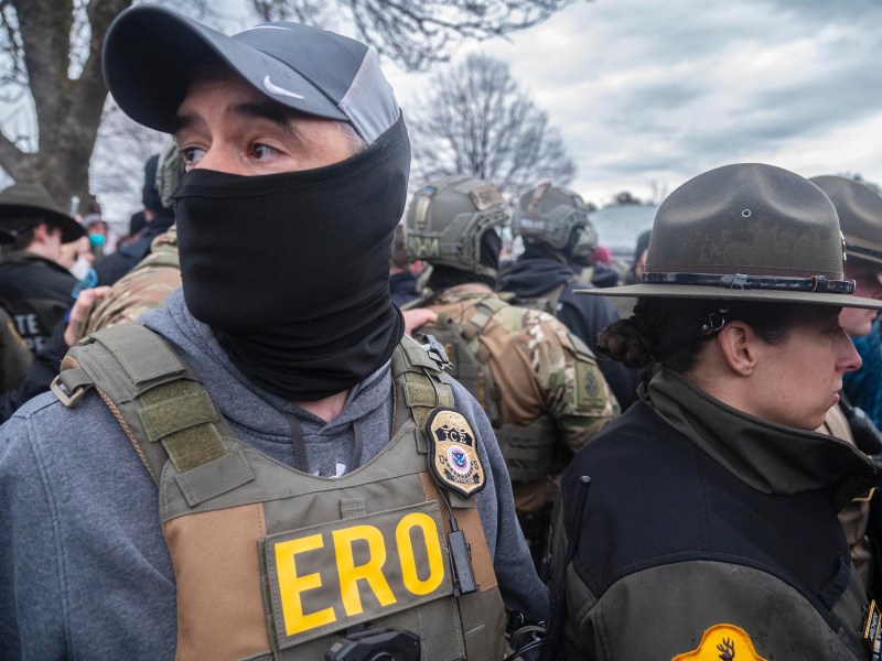 A group of law enforcement officers, some wearing tactical gear and ERO badges, stand closely together outdoors under a cloudy sky.
