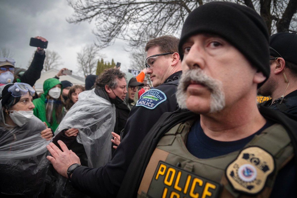 Police officers stand amid a crowd of protesters, some wearing masks and holding plastic sheets, on a cloudy day with bare trees in the background.