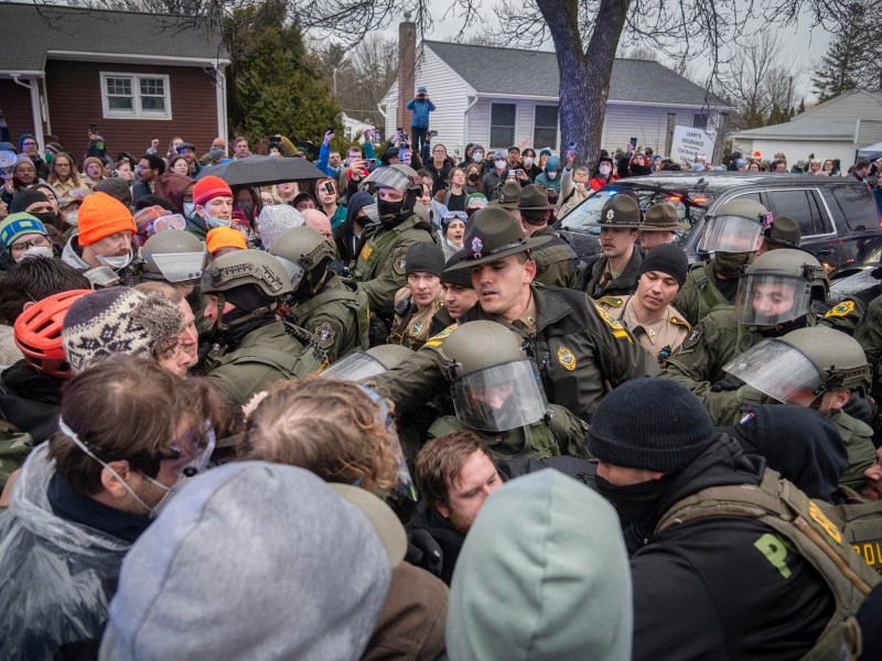 Police officers in riot gear stand in a line facing a dense crowd of protesters on a residential street, with bystanders watching from behind.