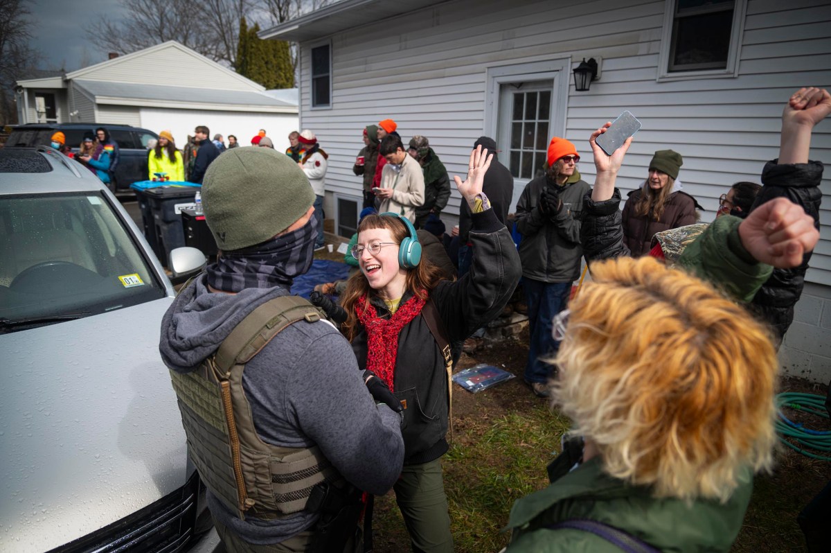 A group of people outdoors near a white house, some raising their arms and wearing headphones or hats, while two people in the foreground interact and smile.