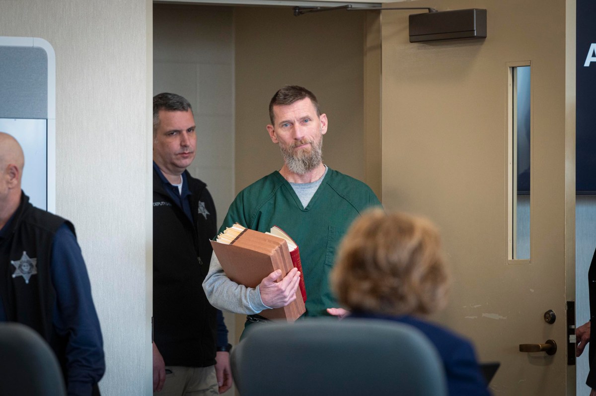 A man in a green prison uniform holds case files while entering a room, accompanied by law enforcement officers.