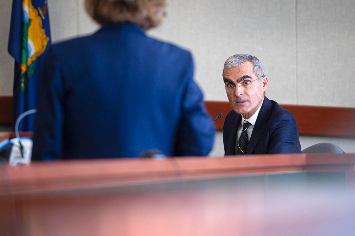 A man in a suit sits at a desk, speaking into a microphone, while a person stands facing him in a courtroom or meeting room.