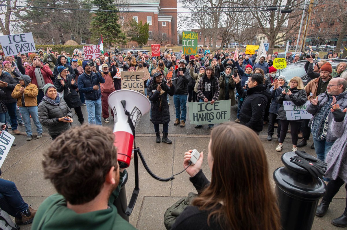 A crowd of people gather outdoors holding signs and banners protesting against ICE, with two speakers addressing them using a megaphone.