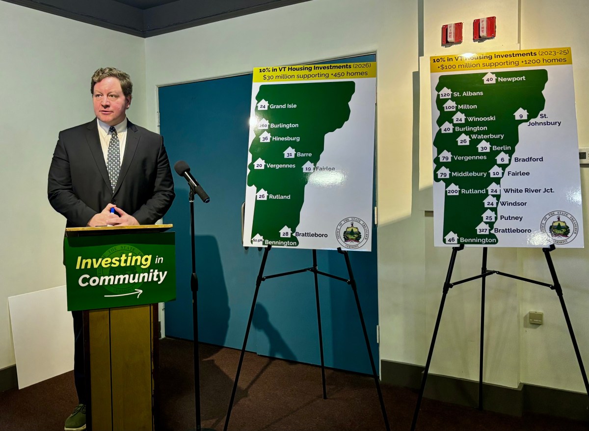 A man in a suit stands at a podium labeled "Investing in Community" next to two posters showing Vermont housing investment locations and funding amounts.