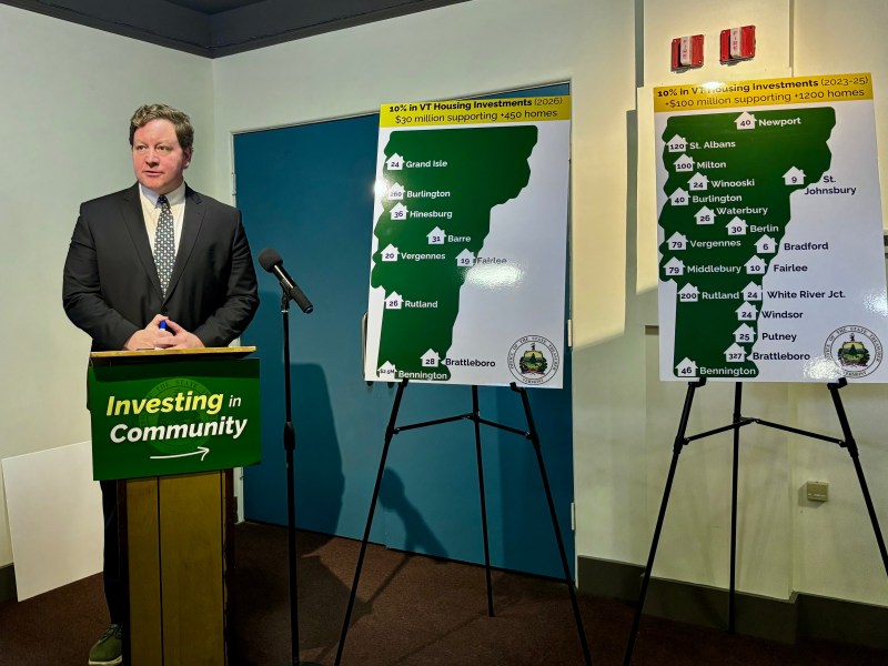 A man in a suit stands at a podium labeled "Investing in Community" next to two posters showing Vermont housing investment locations and funding amounts.
