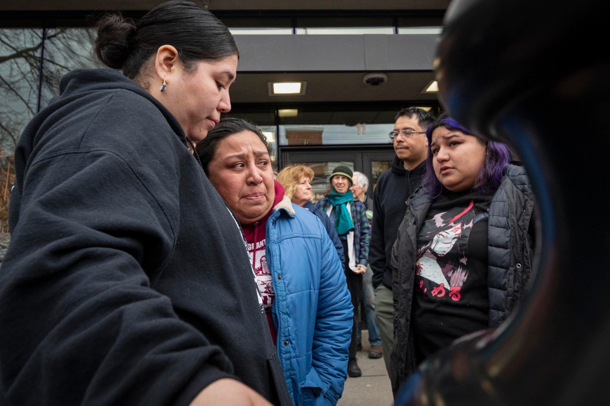 A group of people stand outside a building, some visibly emotional and comforting each other, while others watch in the background.