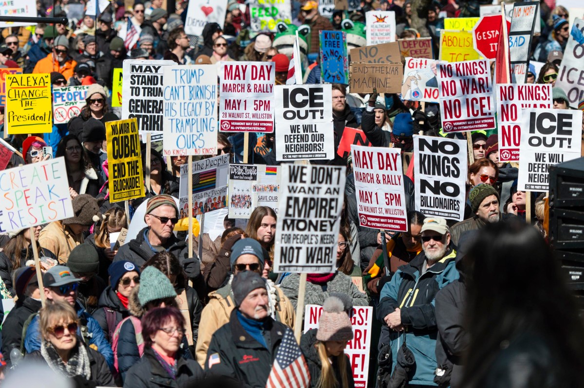 A large crowd of protesters holds various signs, many opposing ICE, Trump, and billionaires, at an outdoor demonstration in daylight.