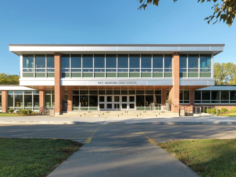 Front view of a modern brick and glass building labeled "Ridge Memorial High School" with large windows, trees in the background, and a sidewalk leading to the entrance.