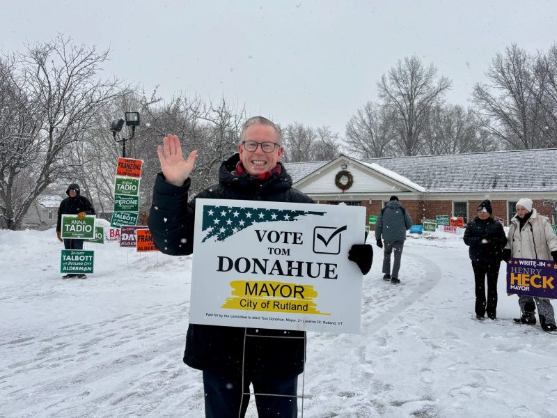 A person stands outside in the snow, smiling and waving, holding a "Vote Tom Donahue Mayor City of Rutland" sign. Other people and campaign signs are visible in the background.