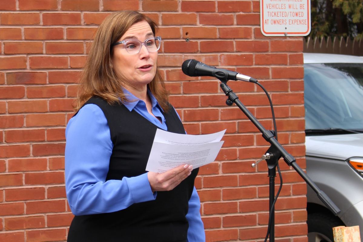 A woman with glasses speaks into a microphone outdoors, holding papers in one hand, standing in front of a brick wall and a parked car.