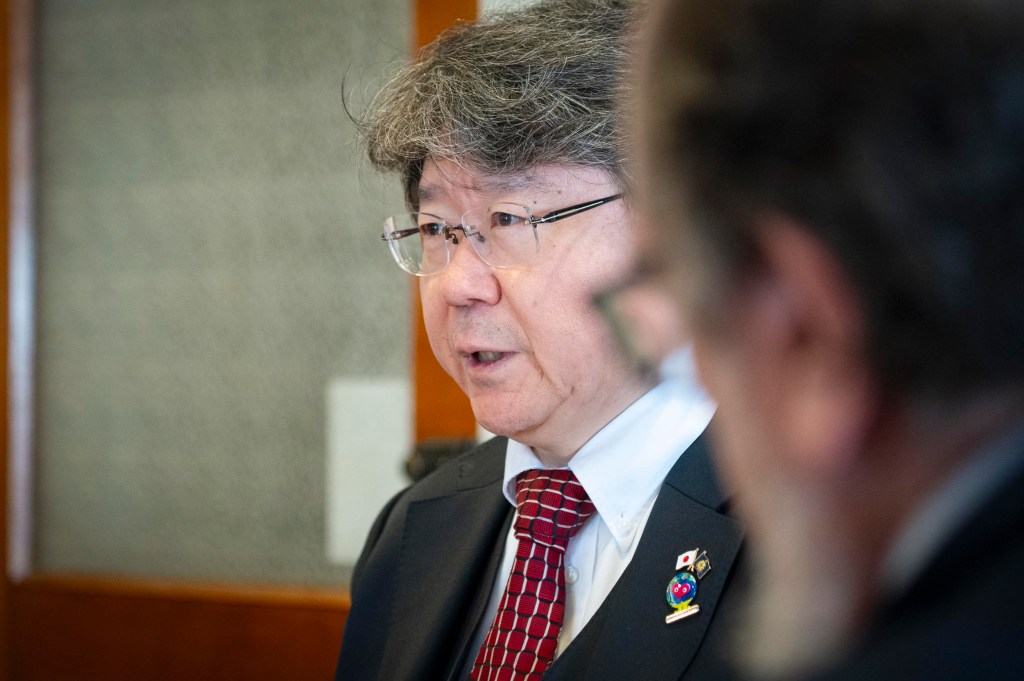 A man in a suit and red patterned tie, wearing glasses, speaks indoors. Another person is visible in the blurred foreground.